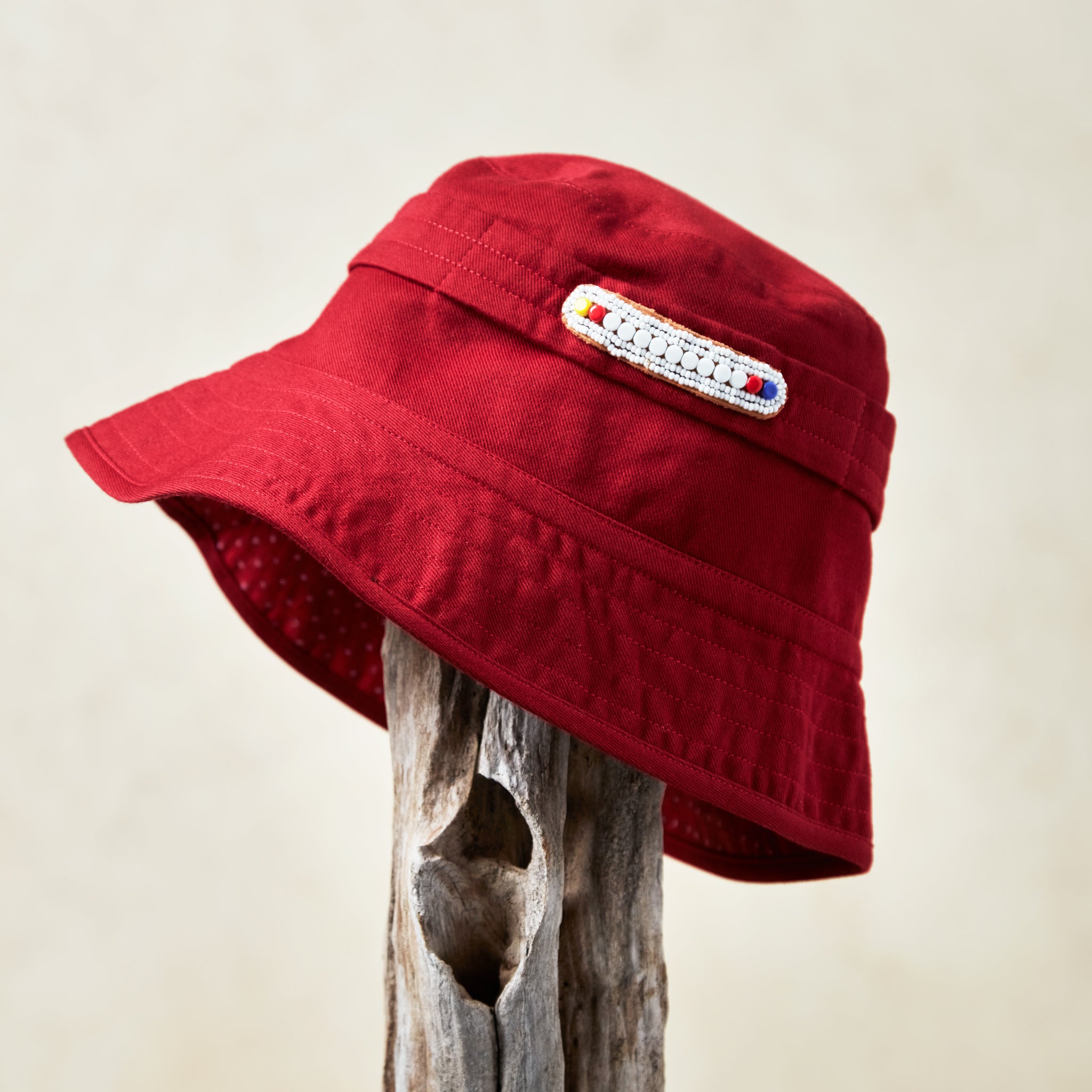 Red bucket hat with a white and colorful beaded patch on a wooden post against a beige background