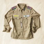Flat front view of Khaki safari shirt with pockets and colorful hand beaded patches on a sand background