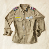 Flat front view of Khaki safari shirt with pockets and colorful hand beaded patches on a sand background