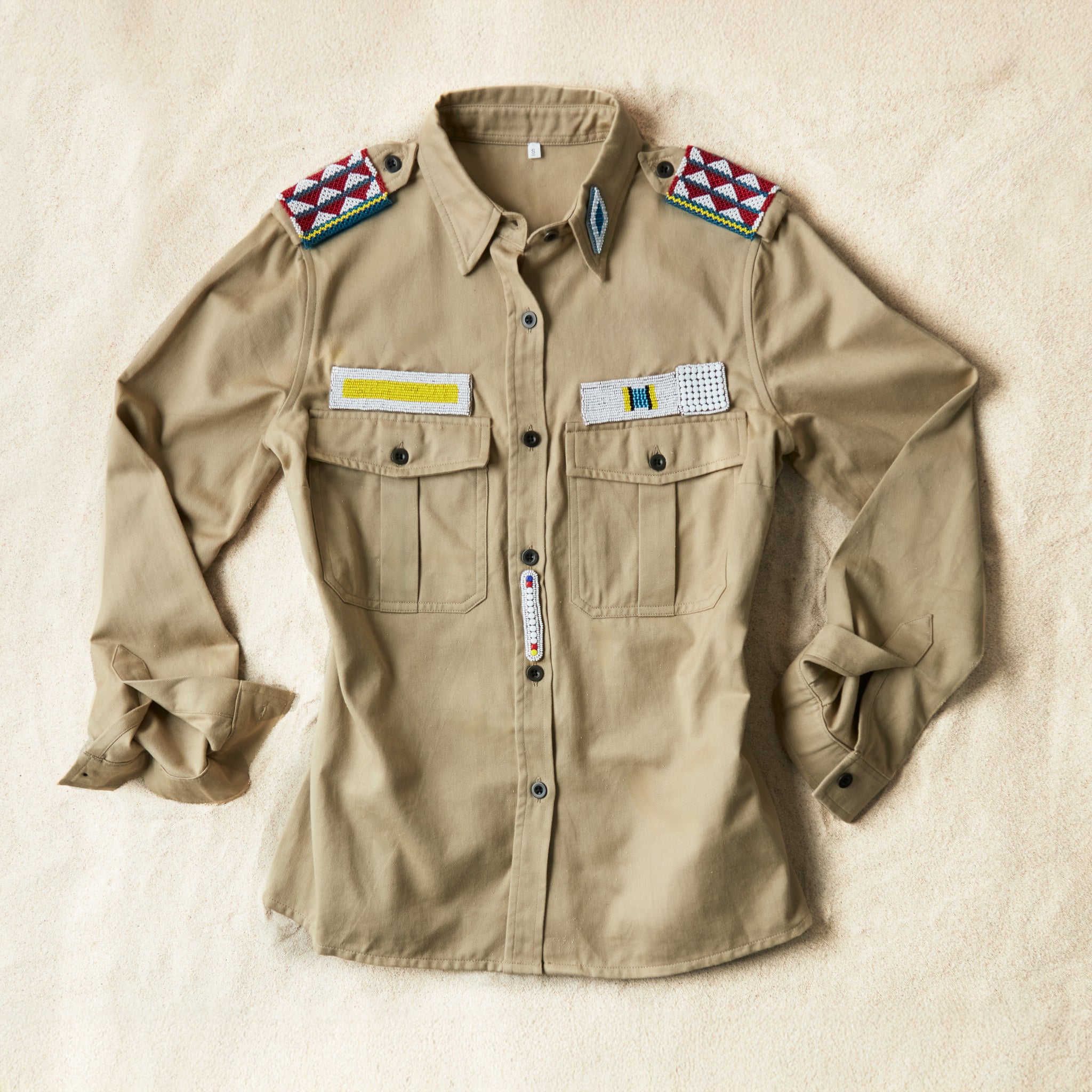 Flat front view of Khaki safari shirt with pockets and colorful hand beaded patches on a sand background