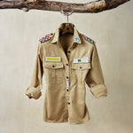 Khaki safari shirt with pockets and colorful hand beaded patches hanging on a wooden branch in front of a beige background