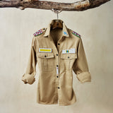 Khaki safari shirt with pockets and colorful hand beaded patches hanging on a wooden branch in front of a beige background