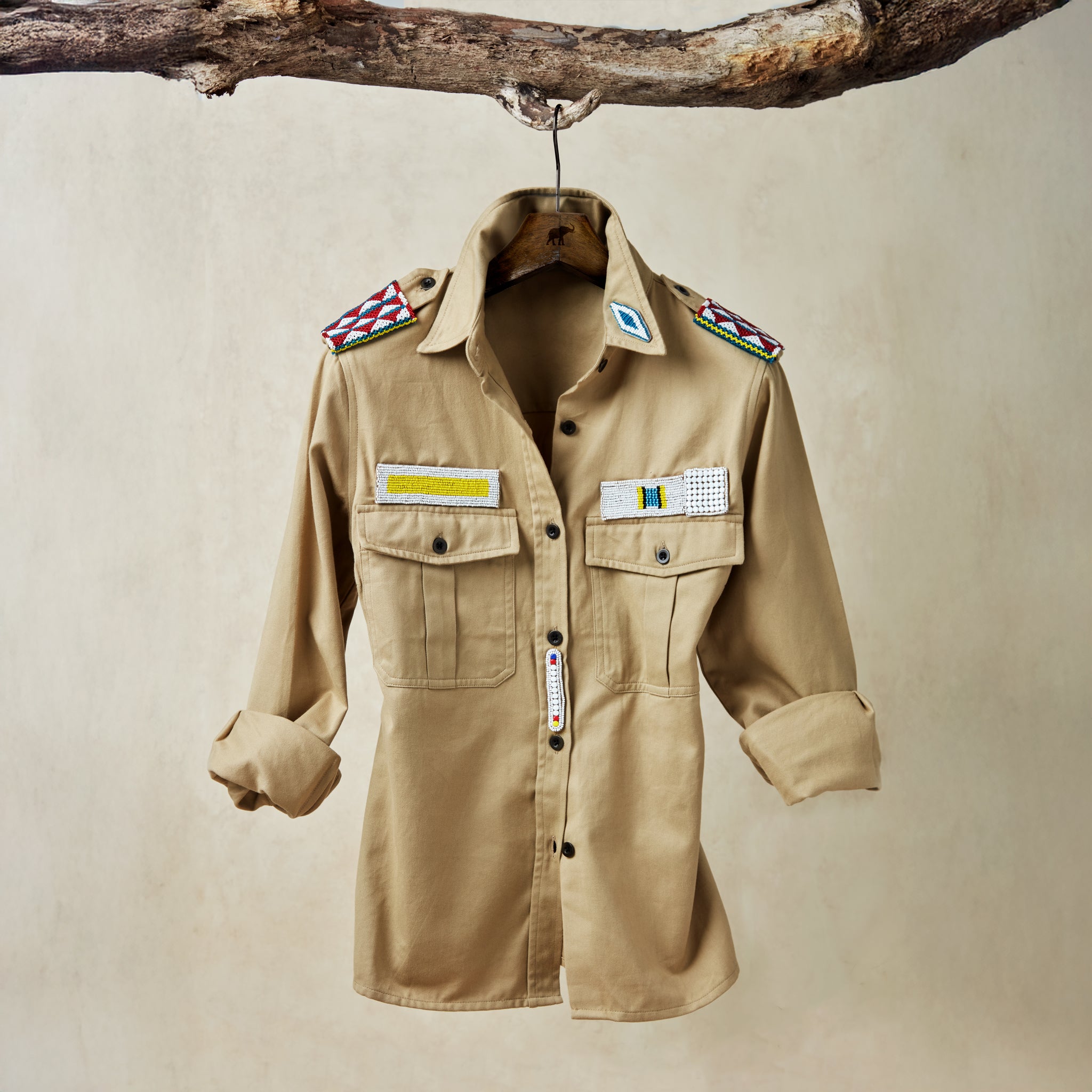 Khaki safari shirt with pockets and colorful hand beaded patches hanging on a wooden branch in front of a beige background
