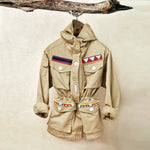 Khaki ranger safari jacket with colorful hand beaded patches hanging on a branch against a beige background