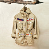 Khaki ranger safari jacket with colorful hand beaded patches hanging on a branch against a beige background