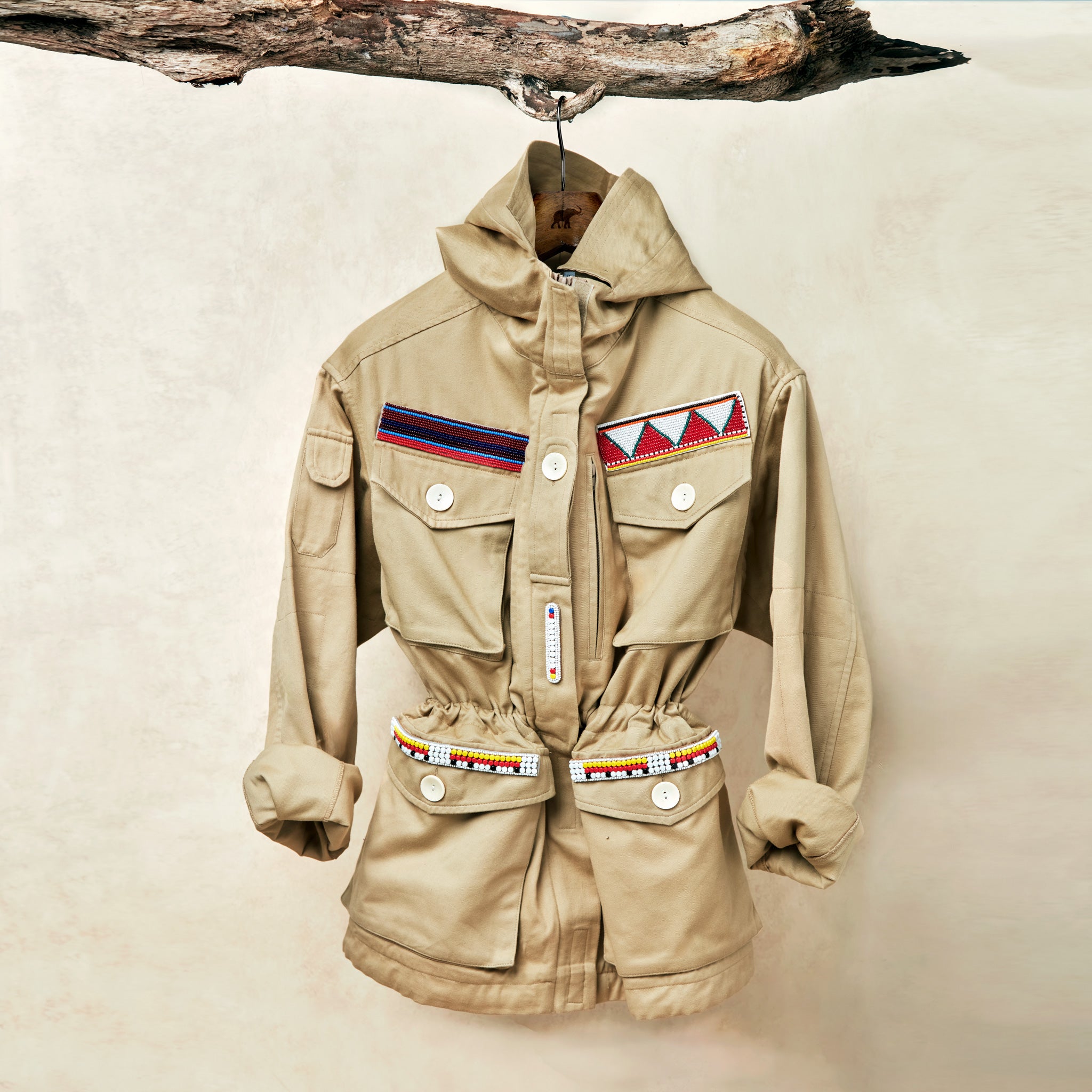 Khaki ranger safari jacket with colorful hand beaded patches hanging on a branch against a beige background