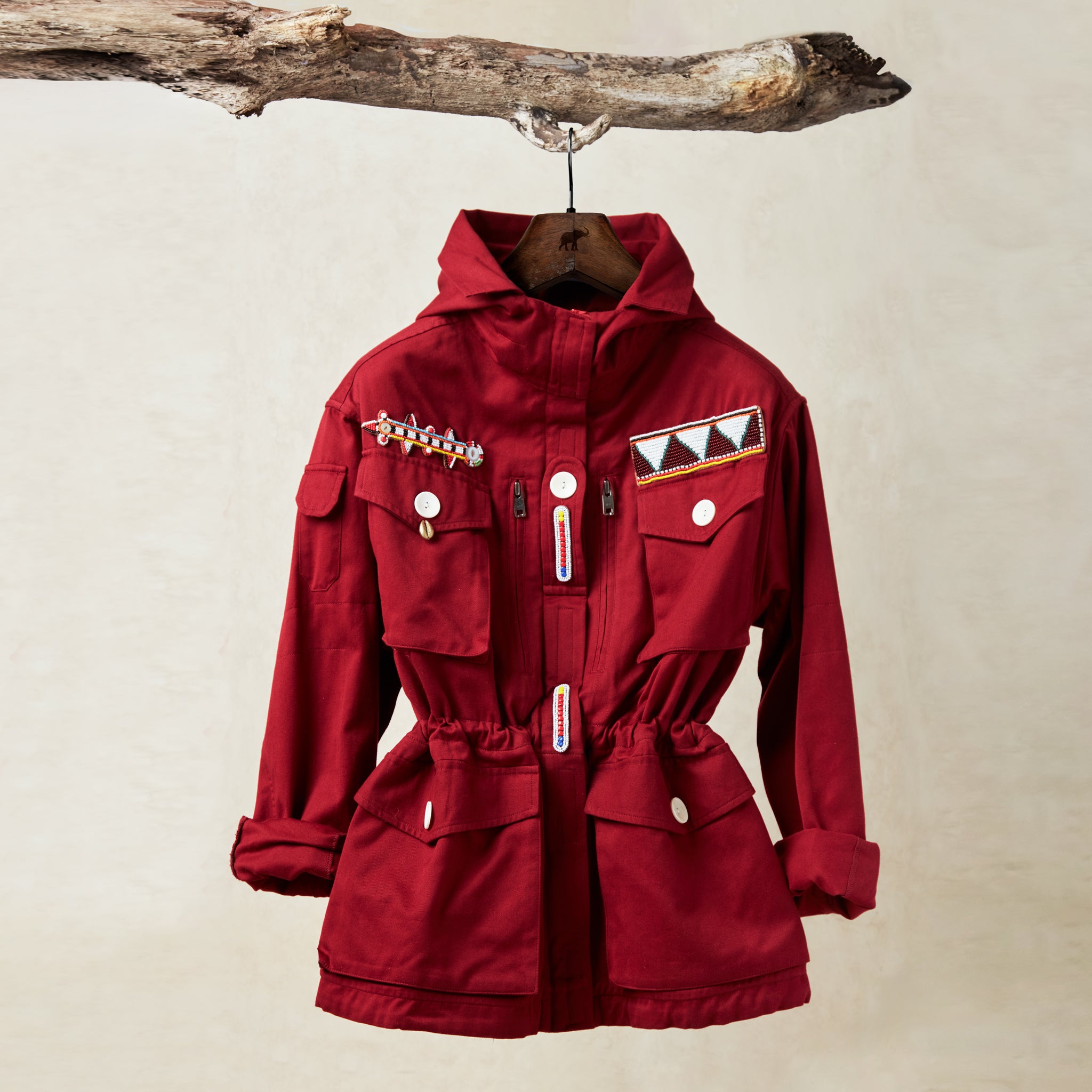 Red ranger safari jacket with colorful hand beaded patches hanging on a branch against a beige background