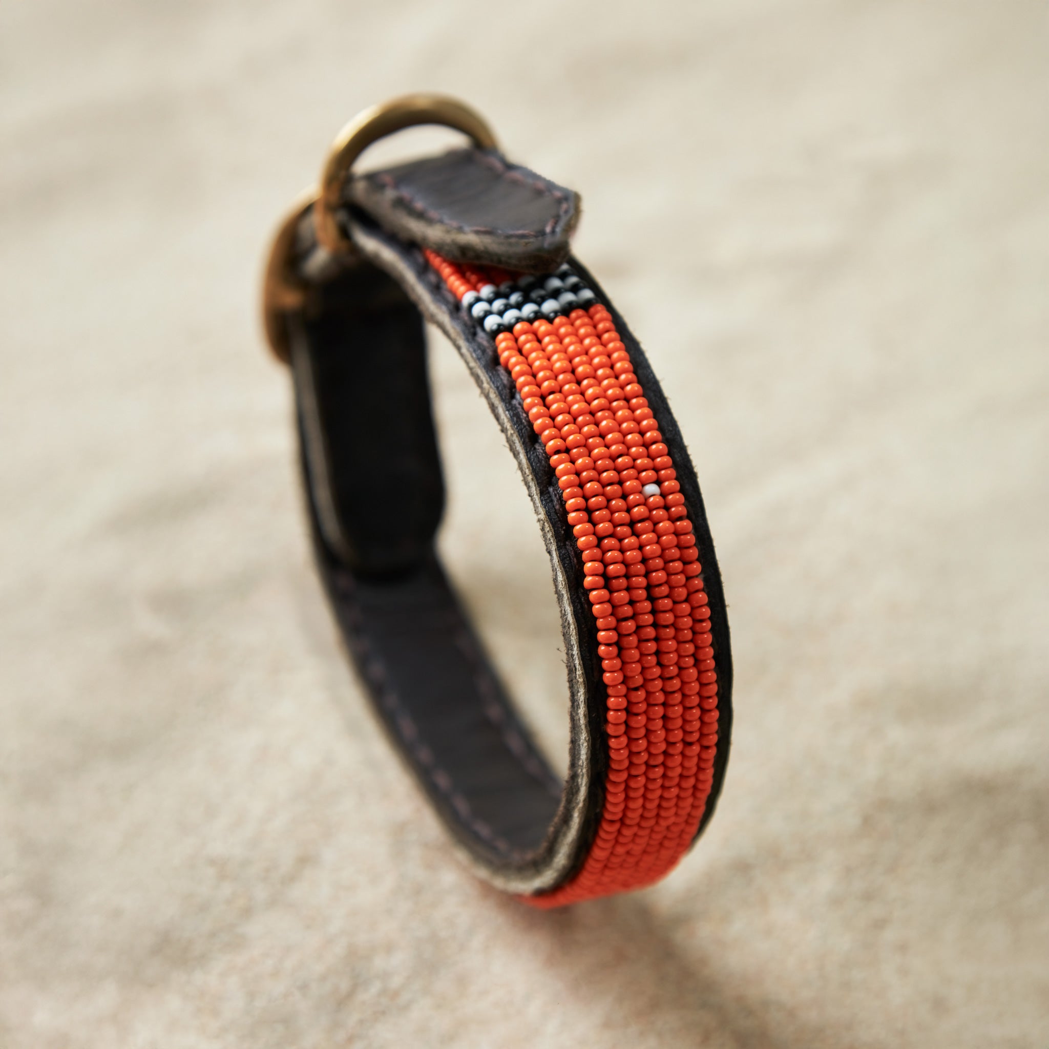 Brown leather dog collar with orange, black, and white beads and a brass buckle on a sand background
