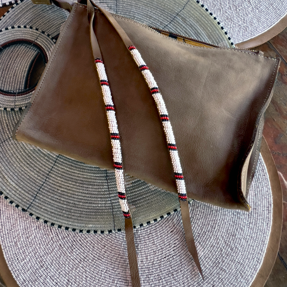 Small sand brown leather pouch with white, black, and red beaded tassel tie laid on top of white and grey beaded neck pieces and products